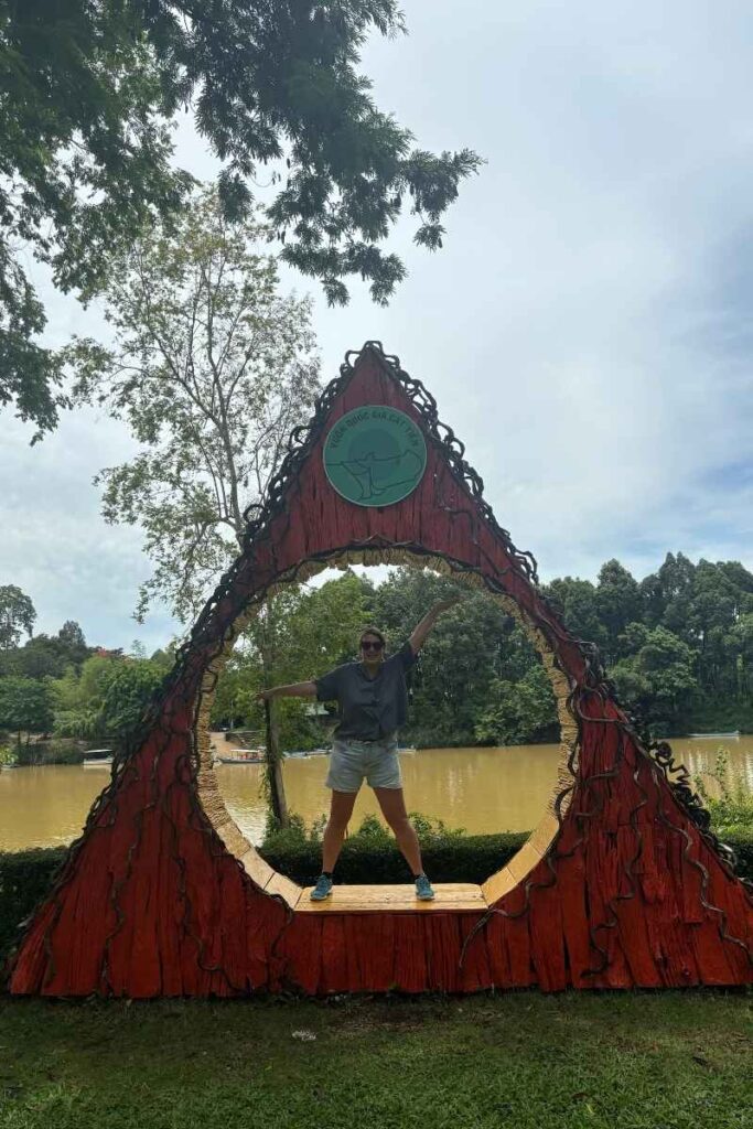Woman posing inside a decorative red archway with a forest and river view behind her at the entrance to Cát Tiên National Park.