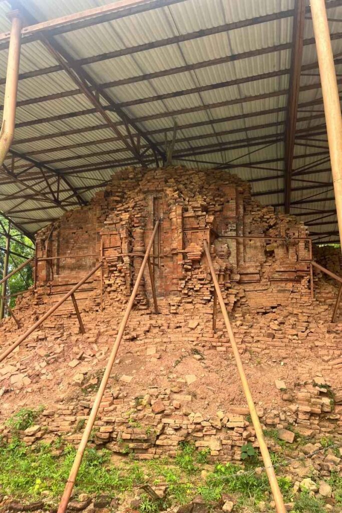 A crumbling temple ruin at My Son Sanctuary protected by a corrugated roof and wooden supports during archaeological restoration.