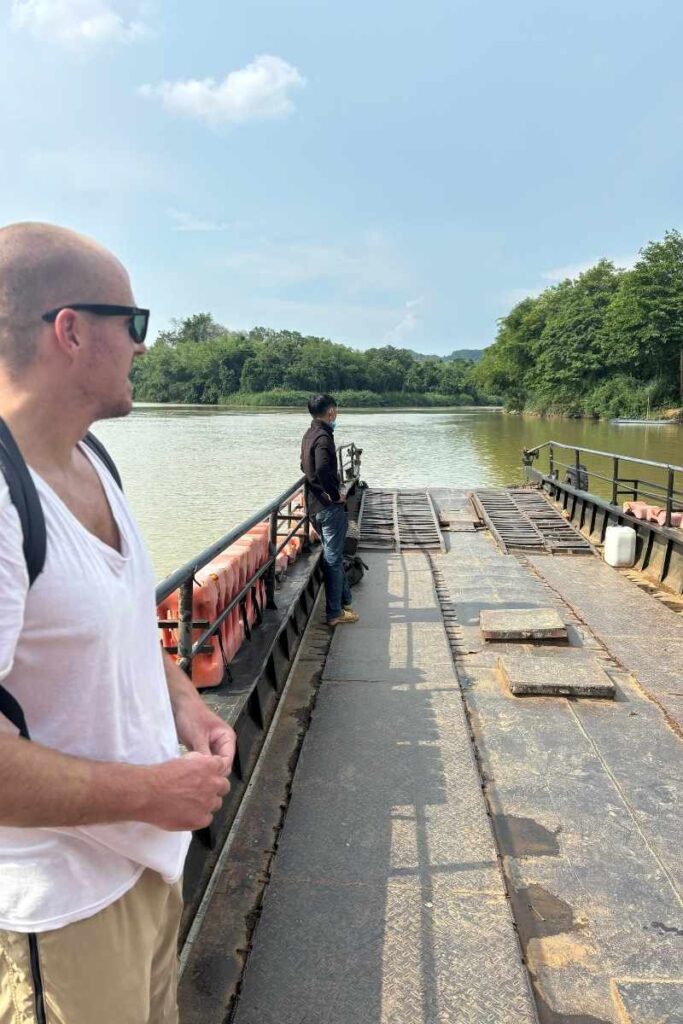 Two men standing on a small ferry crossing the river into Cát Tiên National Park, surrounded by dense green jungle and calm water.