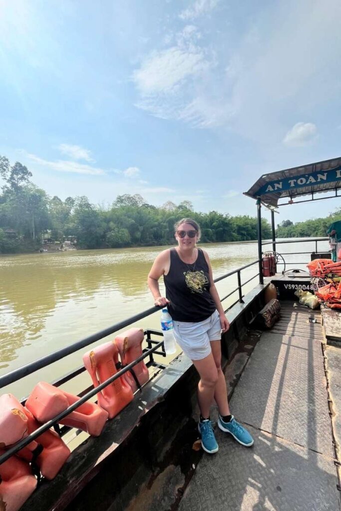 Woman standing on the small ferry crossing the river into Cát Tiên National Park, holding a water bottle under a clear sky.