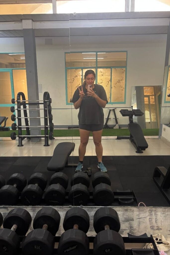 Person in workout clothes stands in front of a large mirror at a hotel gym, flashing a peace sign and holding a phone. The gym is clean and modern with dumbbells, benches, and a rack in view.