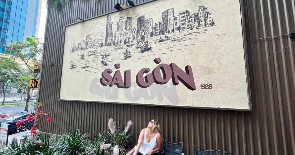 Woman sitting outside a café under a large “Sài Gòn” sign in Ho Chi Minh City, surrounded by tropical plants and casual outdoor seating.