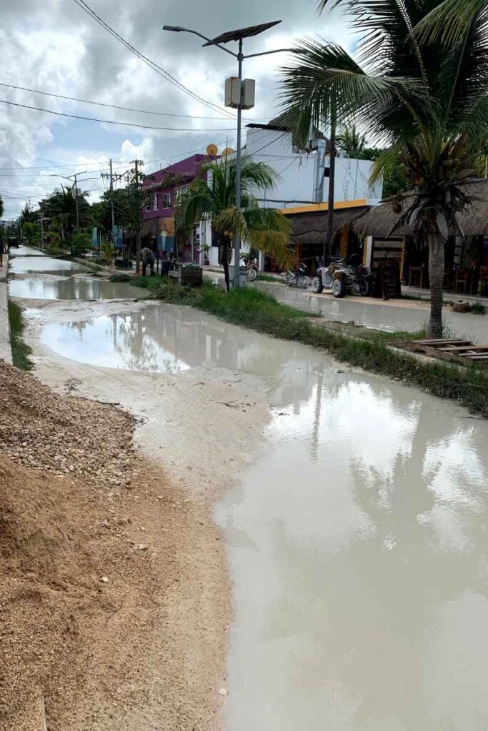 Flooded dirt road in Holbox, Mexico, with murky standing water covering most of the path, reflecting palm trees and nearby buildings. The muddy, impassable terrain highlights the difficulty of travel during rainy conditions, especially without a confirmed hotel.