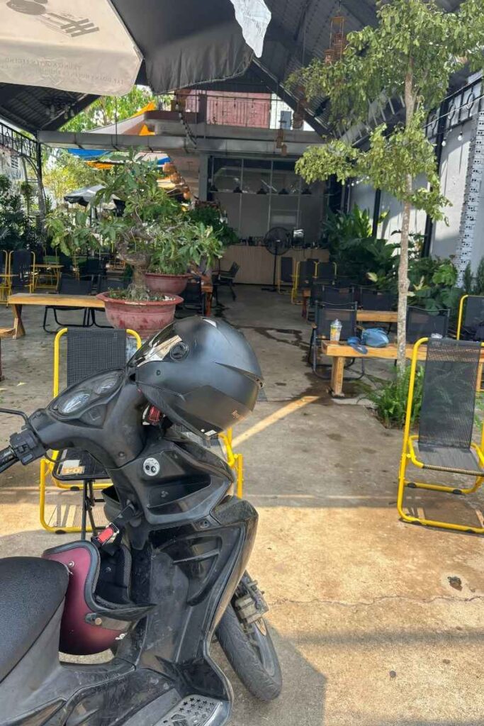 Motorbike with a helmet parked in front of a roadside café with yellow chairs and potted plants, a common pit stop on the way to Cát Tiên Park.