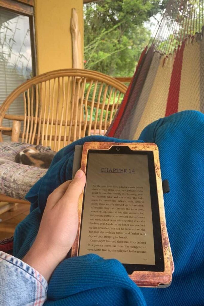 Person lounging in a hammock with a Kindle in hand, open to Chapter 14 of an e-book. A bamboo chair and sleeping dog are visible in the background, capturing a peaceful moment enhanced by the Libby app for free e-book access while traveling.