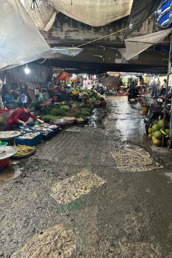 Busy scene inside Cai Be Floating Market with vendors selling fresh produce, fish, herbs, and tropical fruits on low tables and tarps, while customers and motorbikes move through the narrow, wet aisle.