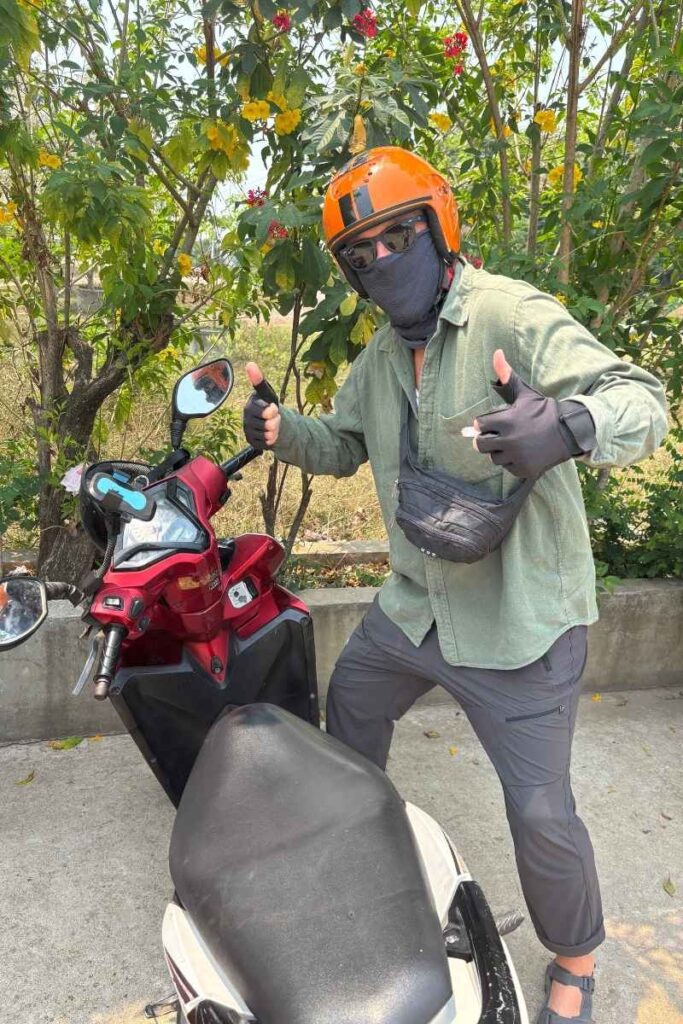 Person wearing an orange helmet, sunglasses, mask, and gloves poses with a red motorbike, giving double thumbs up on a roadside lined with trees. Fully covered for sun and dust protection, this setup reflects a typical motorbike rental experience in Southeast Asia using the best travel apps for navigation and bookings.