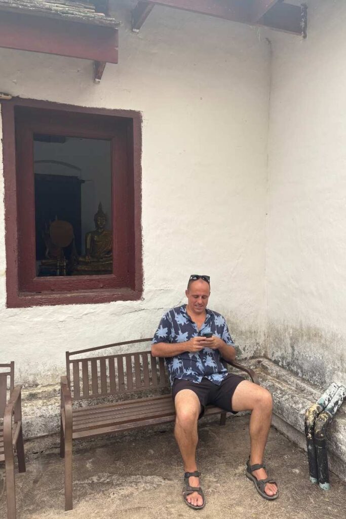 Person sitting on a wooden bench outside a weathered white temple wall, wearing a blue floral shirt and looking down at their phone. A red-framed window reveals golden Buddha statues inside, suggesting a quiet moment of travel planning or checking the best travel apps while exploring a cultural site.