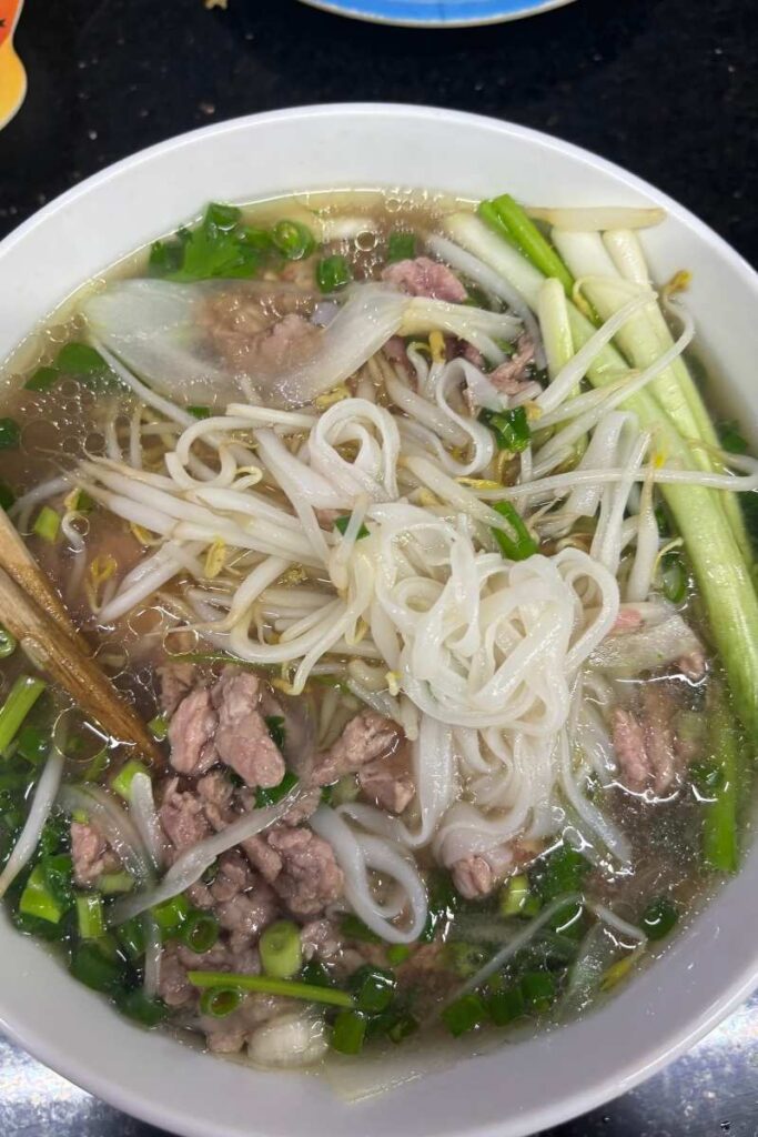 Close-up of a steaming bowl of pho with rare beef slices, rice noodles, scallions, and bean sprouts, a must-try dish when visiting HCMC.