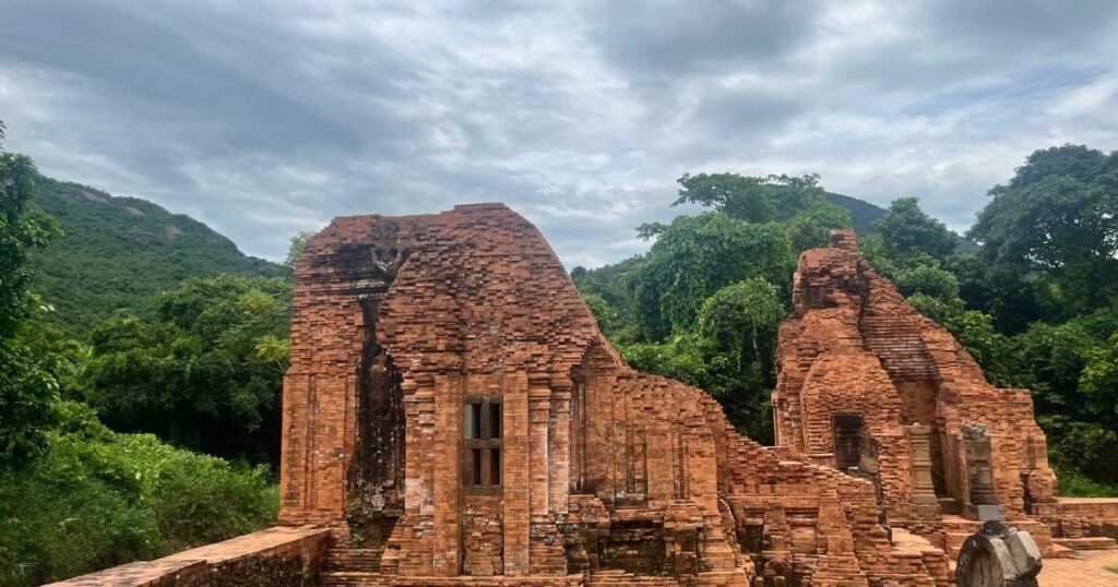 A wide-angle view of partially restored Cham temple towers at My Son, with reddish brick walls rising out of a green jungle setting beneath a cloudy sky.