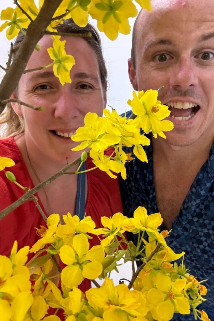 Close-up of two people smiling behind a branch of vibrant yellow hoa mai flowers, a traditional Tet decoration in Ho Chi Minh City.