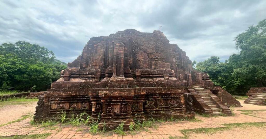 Wide view of a prominent Cham temple at My Son, featuring intricate brickwork, steps, and decorative carvings, framed by dense trees