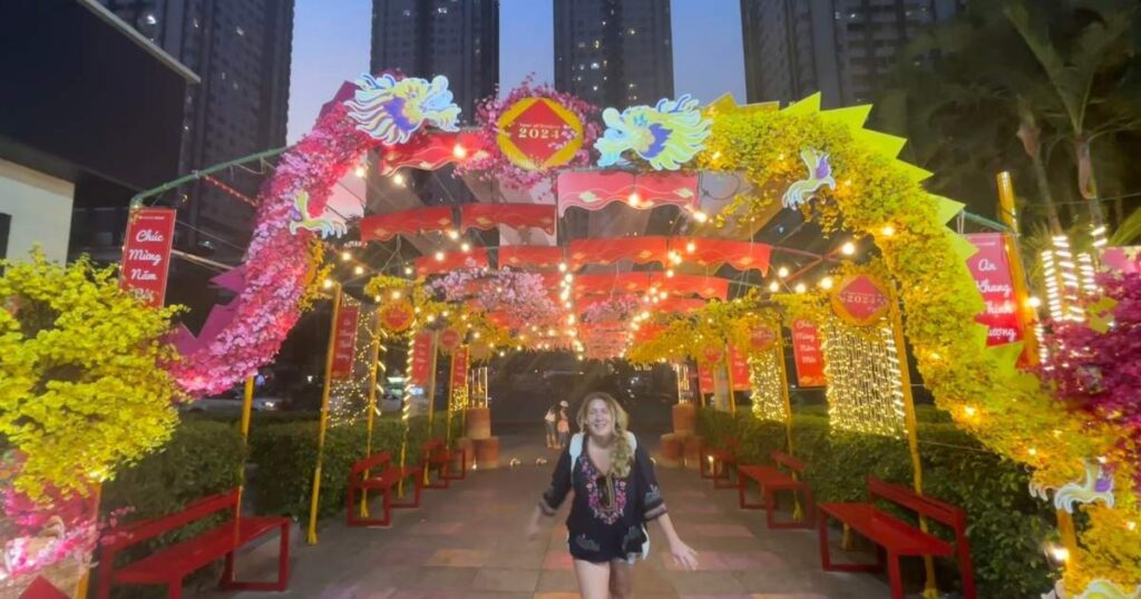 Tet celebration walkway in Saigon decorated with red lanterns, yellow flowers, and banners for the 2024 Lunar New Year, with a smiling visitor walking through the festive archway.