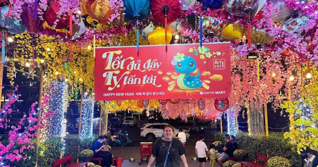 Colorful Tet display at Lotte Mart in Ho Chi Minh City featuring lanterns, cherry blossoms, and a 2025 Year of the Dragon sign reading "Tết đã đầy Tý tấn tài" above smiling visitors.
