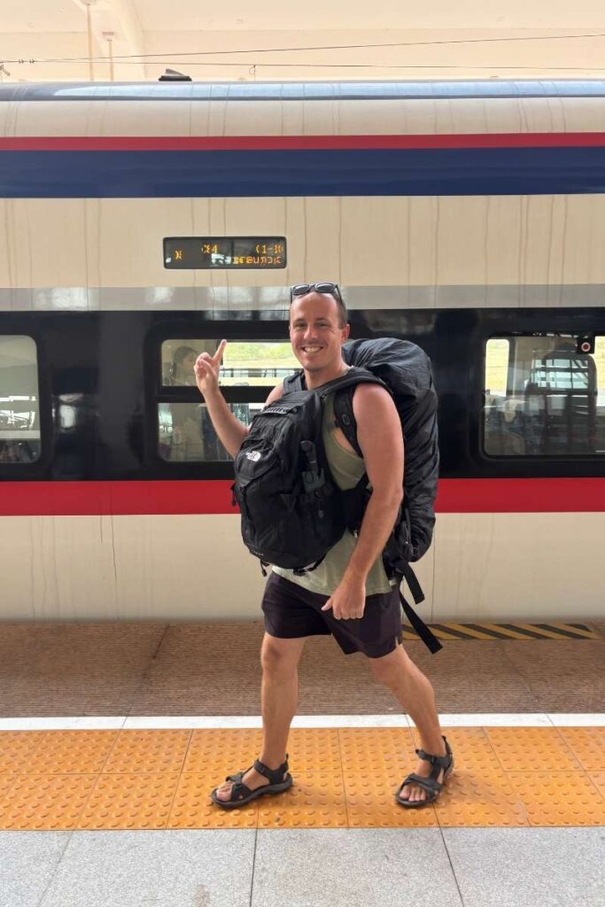 Smiling traveler with a large black backpack stands in front of a high-speed train, pointing up at the digital sign above the platform.