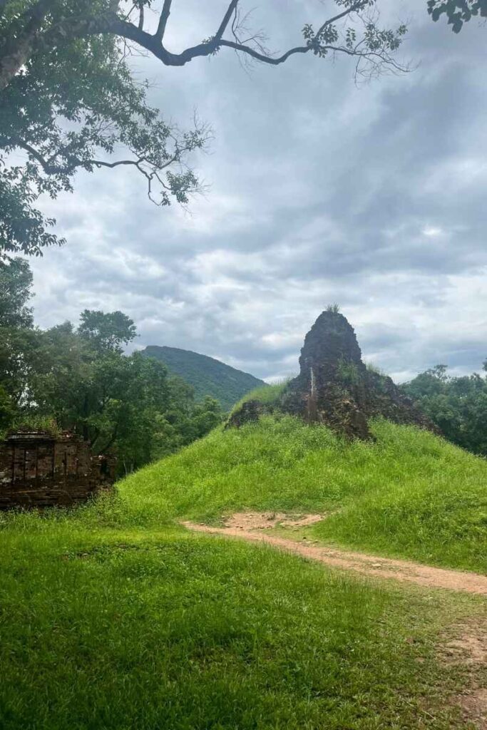 A partially collapsed Cham tower covered in vegetation rises from a grassy mound at My Son Sanctuary, framed by jungle and mountains.