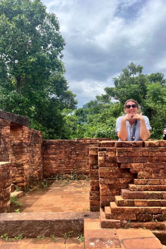 A woman leans on a brick wall, smiling with her chin in her hands, surrounded by Cham ruins and lush greenery.