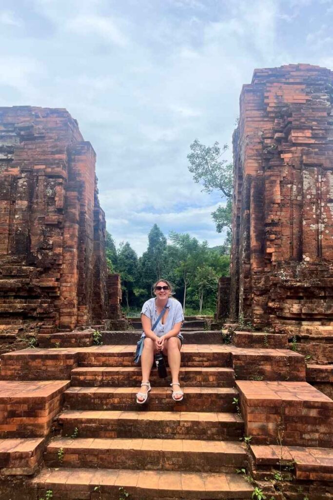 A woman sits on the brick steps between two crumbling temple towers at My Son Sanctuary, surrounded by green jungle and historic red ruins.