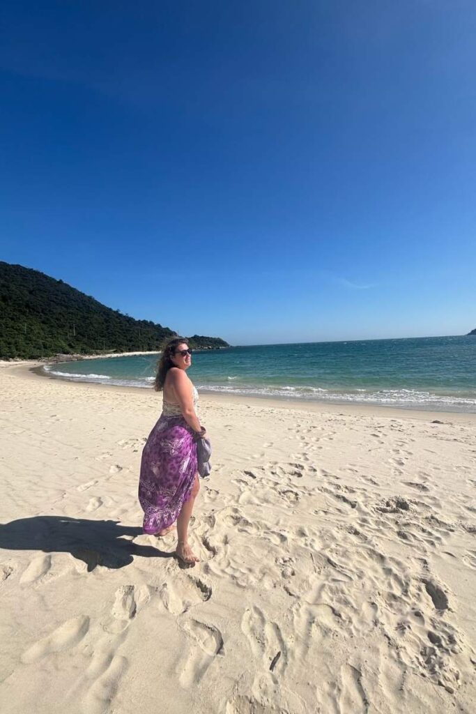 Woman walking along the soft white sand at Bai Ong Beach on Cham Island, with turquoise water and a forested hill in the distance.