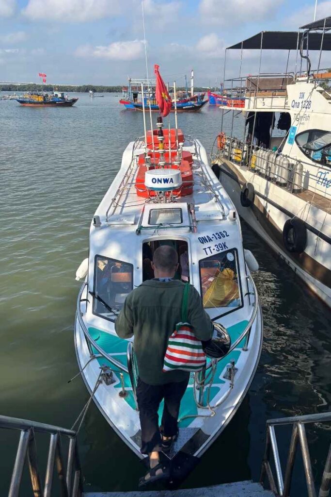 Man stepping onto a silver speedboat ferry at the dock, preparing to travel to Cham Islands, with other colorful boats in the background.