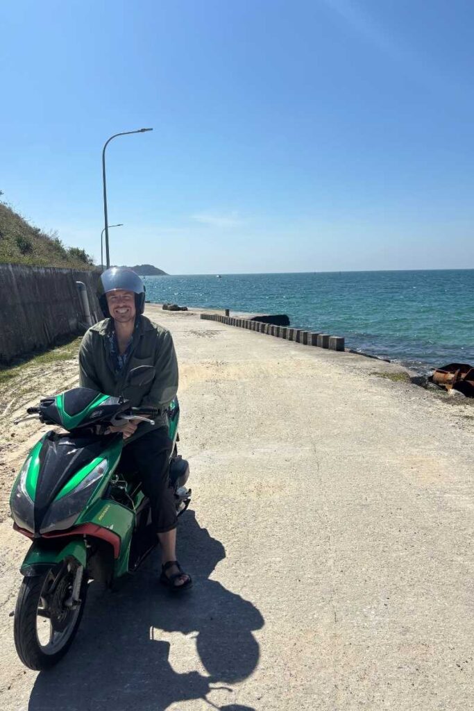 Man sitting on a green motorbike along a quiet coastal road on Cham Island Vietnam, with the ocean stretching out beside her under a bright blue sky.