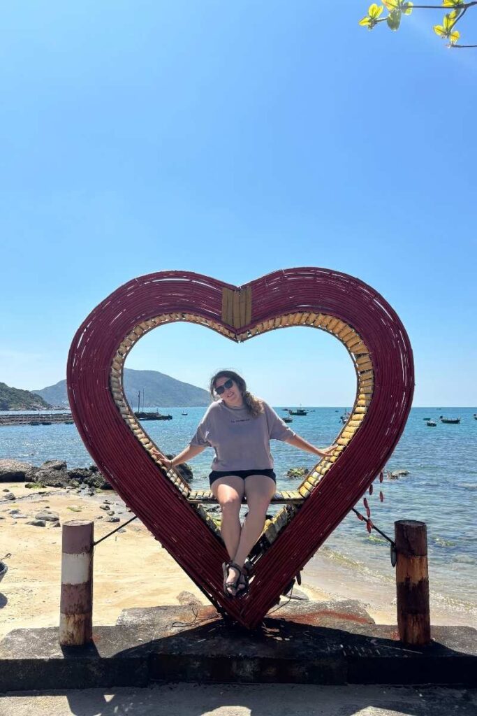 Woman sitting on a heart-shaped swing by the water in Bai Huong village on Cham islands vietnam, with boats and blue sky in the background.