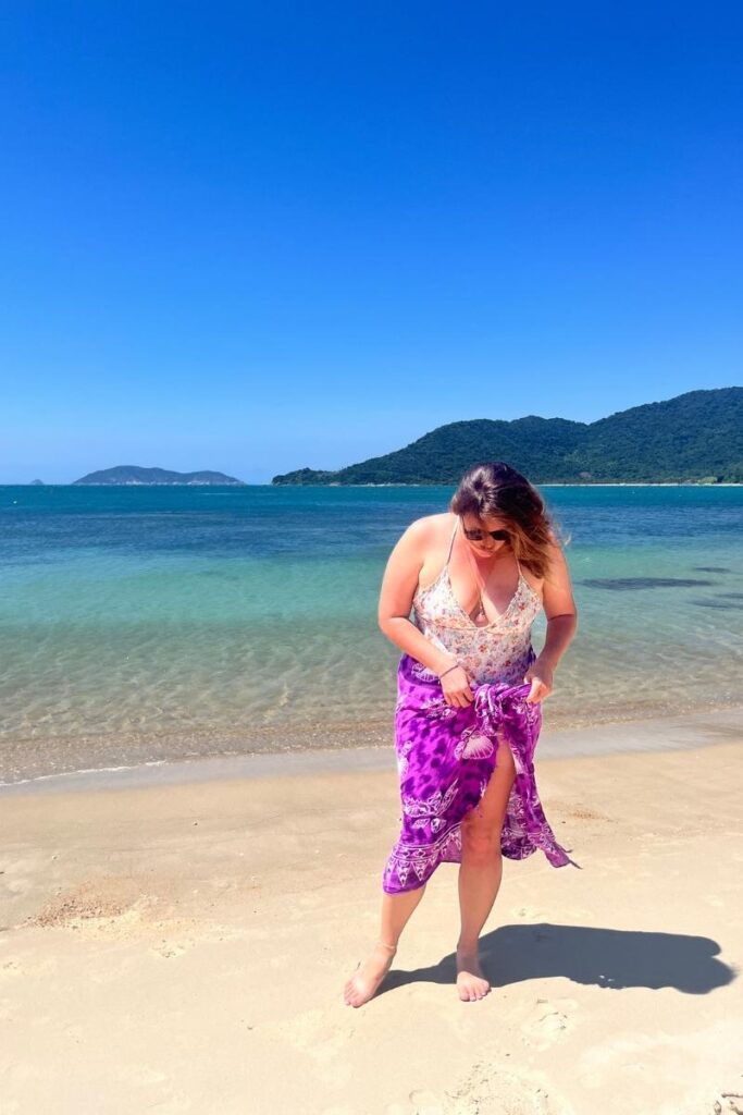Woman standing on the soft sand of Cham island beaches, adjusting her sarong with clear turquoise water and lush hills in the background.