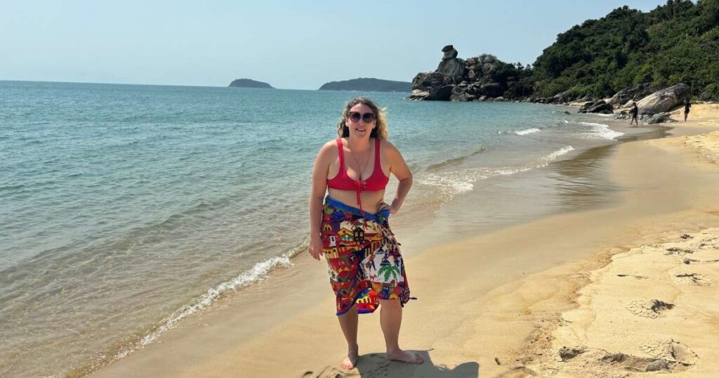 Woman smiling on a quiet beach on Cu Lao Cham after lunch during a boat tour, with clear water, rocky cliffs, and forested hills in the background.