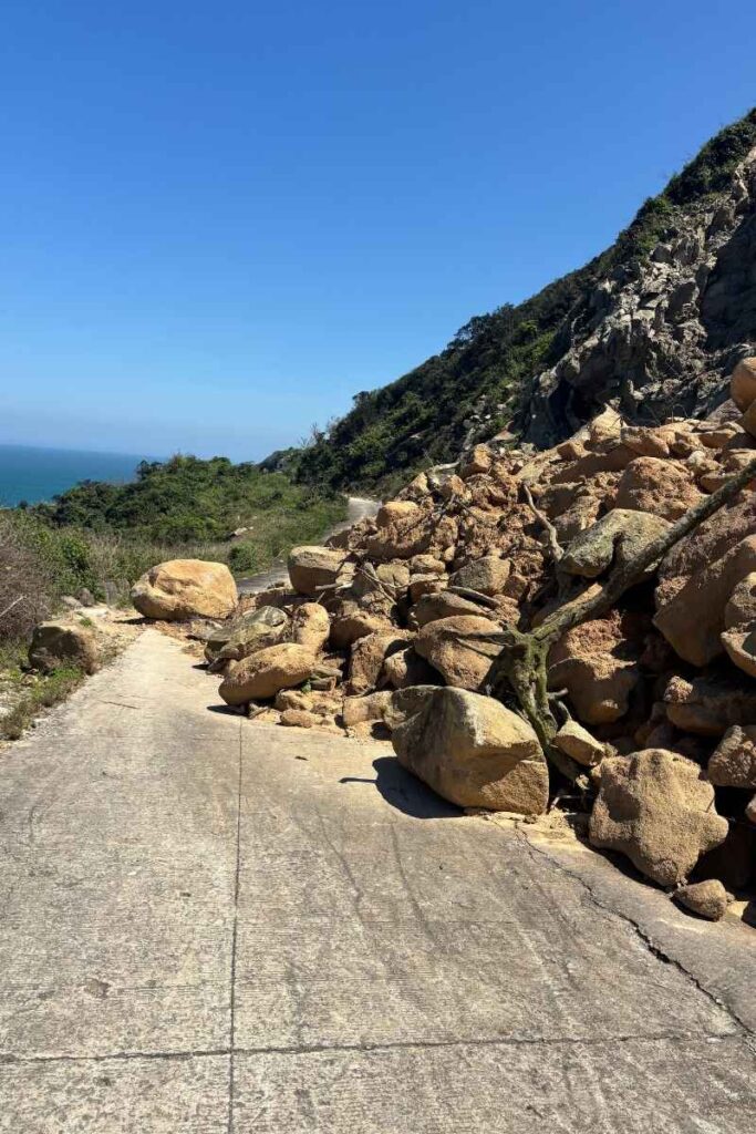 Large rockslide blocking a narrow concrete road on Cu Lao Cham, with scattered boulders piled against a hillside and ocean visible in the distance.