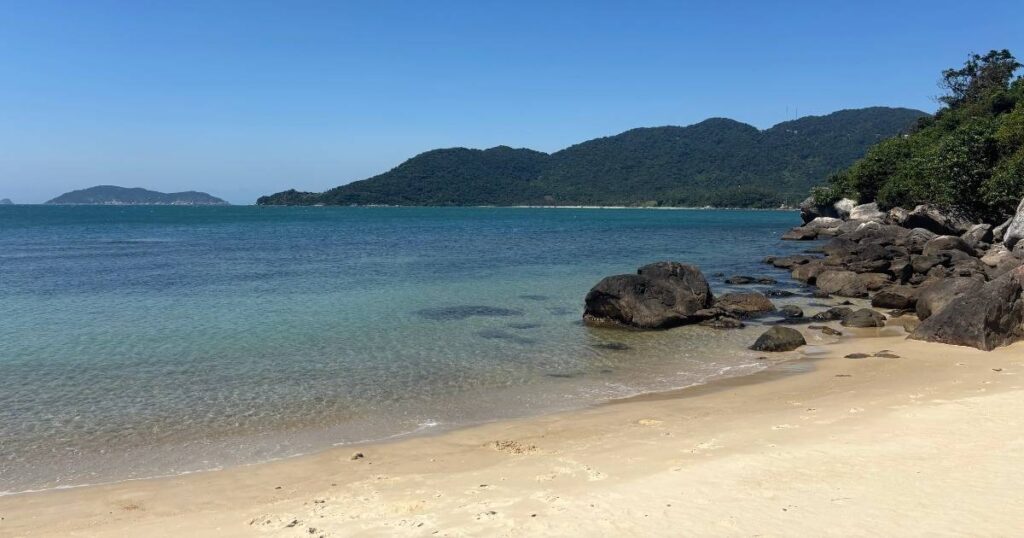 Peaceful beach on Cham Island with soft golden sand, clear turquoise water, and smooth boulders lining the shoreline beneath forested hills.