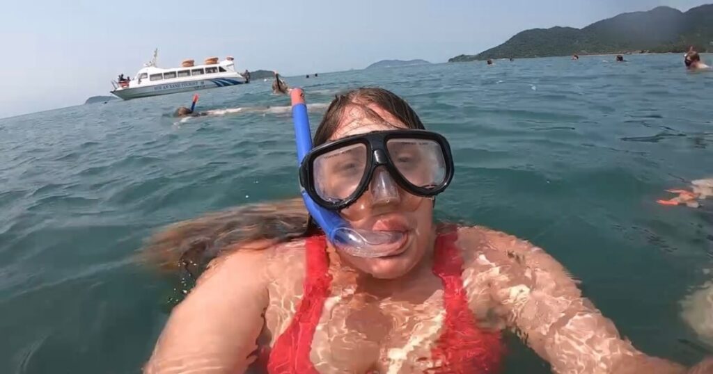 Woman in a red swimsuit wearing a snorkel mask during a boat tour near Cham Island UNESCO Biosphere Reserve, with others swimming nearby and a tour boat in the background.