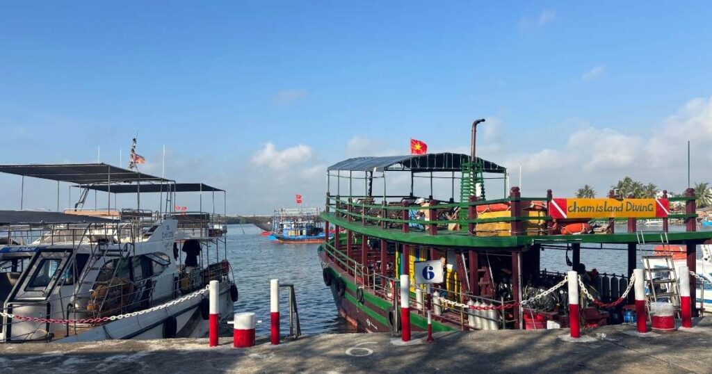 Dock with several boats including a green and red one labeled "Cham Island Diving," where visitors board speedboats to reach Cham Island under a clear blue sky.