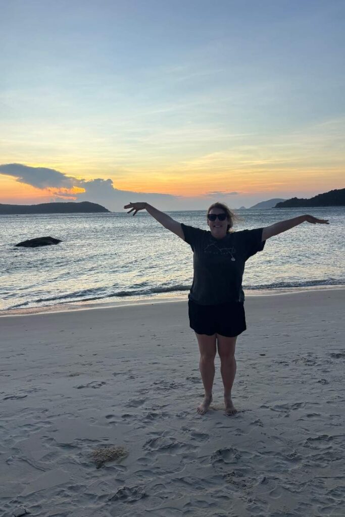 Woman standing with arms outstretched on Bai Xep beach during sunset on Cu Lao Cham, with soft waves and glowing sky behind her.