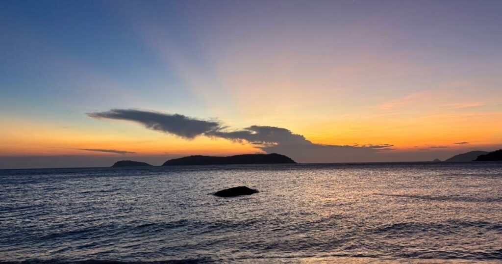Golden and purple hues stretch across the sky as the sun sets behind the islets near Cham Island, with gentle waves and a single rock in the foreground.