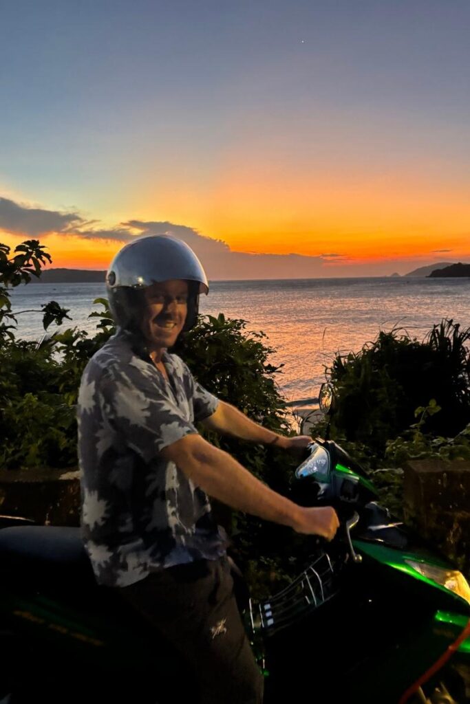 Man on a motorbike smiling during sunset on Cham Island, with vivid orange and purple hues over the sea in the background.