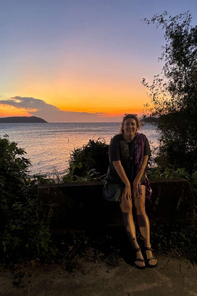 Woman smiling in front of a vibrant Cham islands vietnam sunset, with orange and purple skies above the ocean and silhouetted greenery around her.