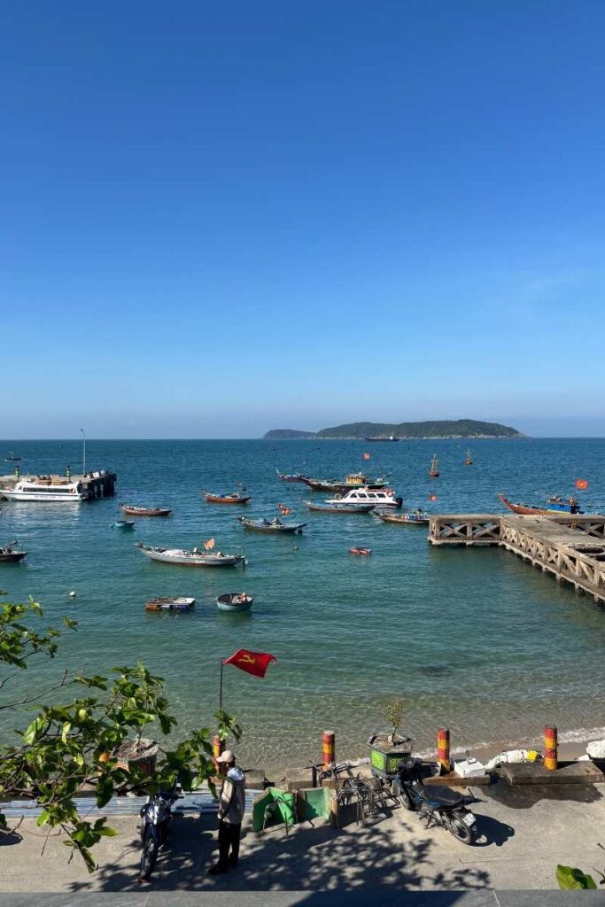 Ocean view from Tuan Thanh Homestay on Cham Island, overlooking a small dock, anchored boats, and a distant islet under a clear blue sky.