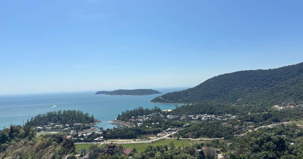 Viewpoint overlooking a village on Cham Islands, with green hills, clustered homes, and calm blue ocean stretching out toward offshore islets under a bright sky.