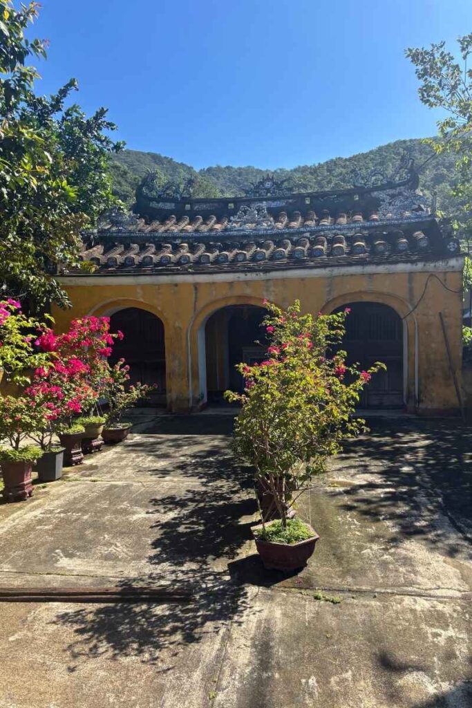 A weathered yellow temple on Cu Lao Cham with arched doorways, potted plants, and cracked concrete surrounded by green hills.